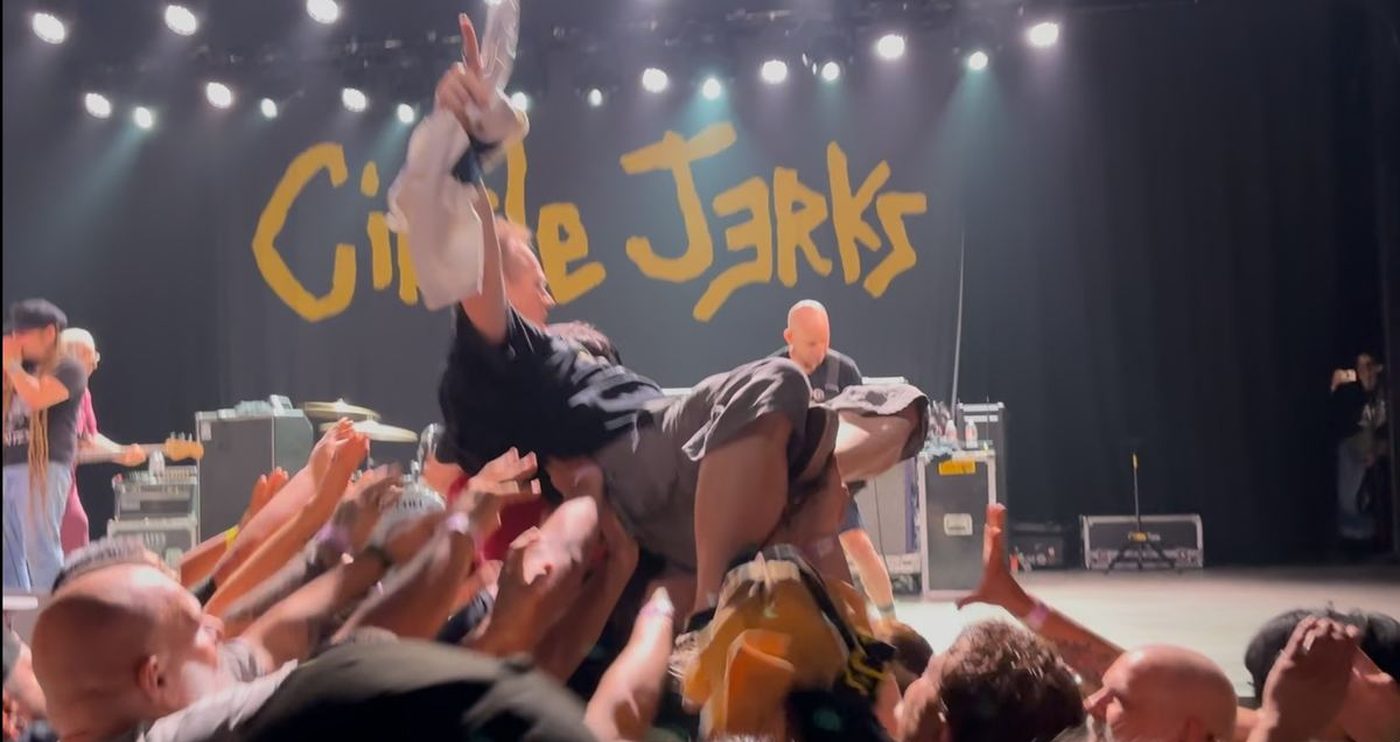 A person carried horizontally on the raised hands of a crowd at a punk show, band backdrop behind them